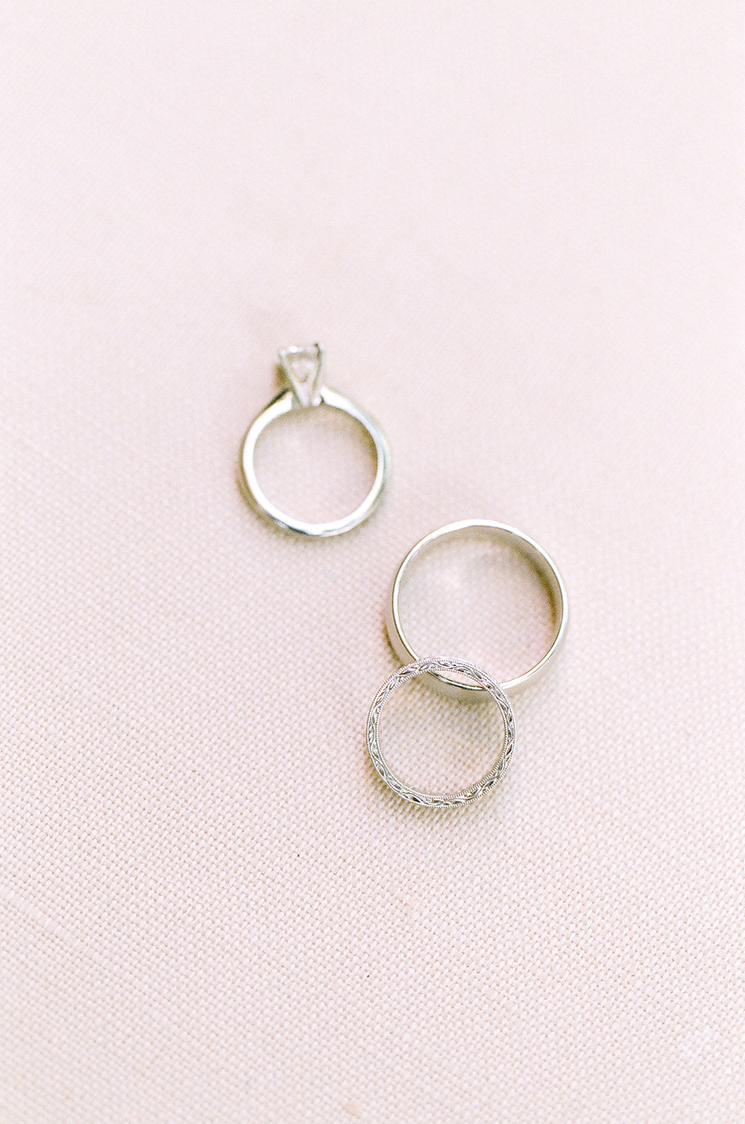photo of two wedding bands and an engagement ring sitting on top of a pale pink background