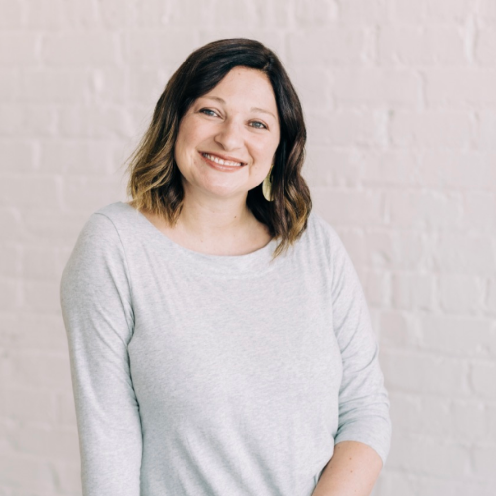 Photo of a woman (Lanna Britt) with brown shlder length hair, in a light grey long sleeve shirt, smiling at the camera in front of a white stone wall