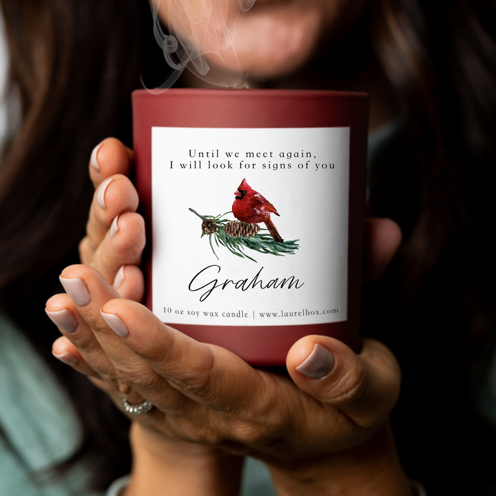 A woman holding a red-coated candle with a white sticker including the text Until we meet again, I will look for signs of you, Graham, with a cardinal image. 
