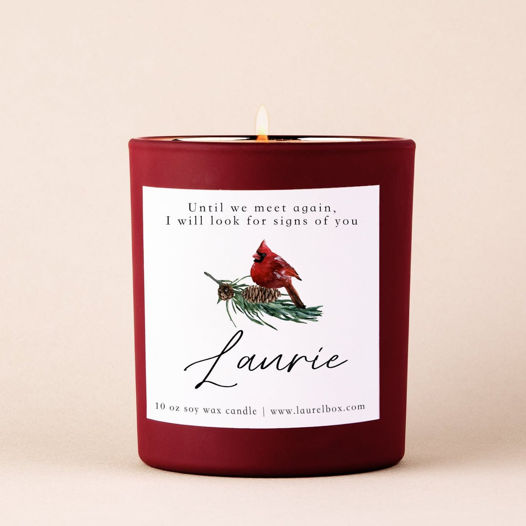 A red-coated candle with a white sticker including the text Until we meet again, I will look for signs of you, Laurie, with a cardinal image against a tan background