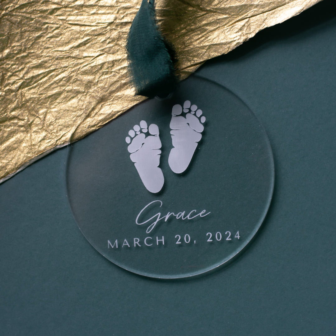 This is a close-up of a personalized acrylic glass ornament with a baby footprint, the text "Grace" and "March 20, 2024" engraved, and a dark green ribbon at the top. The background is a teal surface with a gold fabric to the left.