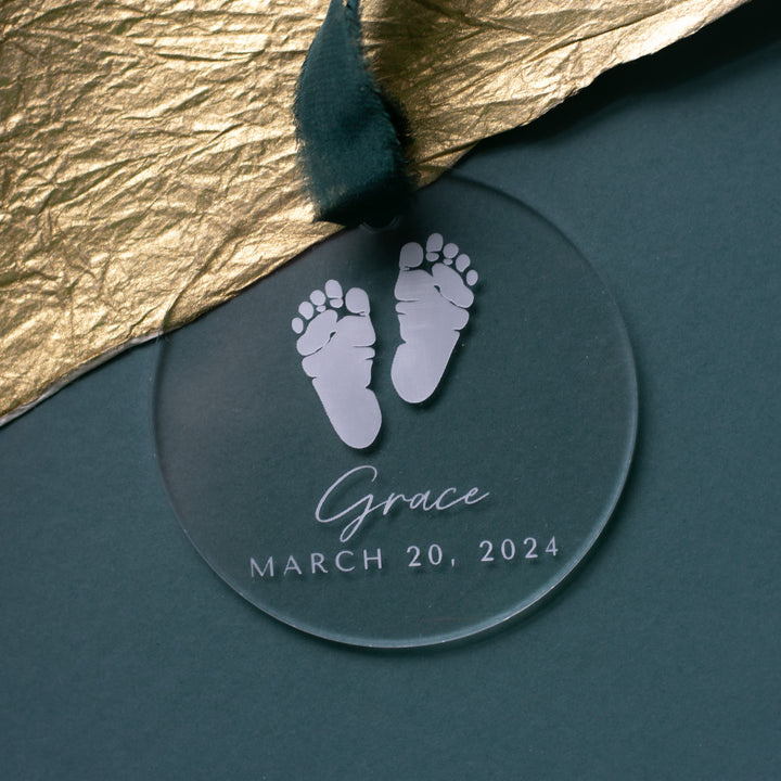 This is a close-up of a personalized acrylic glass ornament with a baby footprint, the text "Grace" and "March 20, 2024" engraved, and a dark green ribbon at the top. The background is a teal surface with a gold fabric to the left.