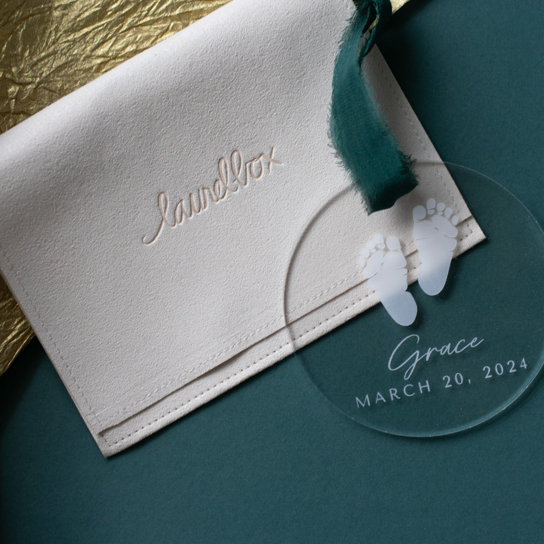 A close-up of a personalized acrylic glass ornament with a baby footprint, the text "Grace" and "March 20, 2024" engraved, and a dark green ribbon at the top resting on a tan suede pouch with laurelbox engraved. The background is a teal surface with a gold fabric to the left.