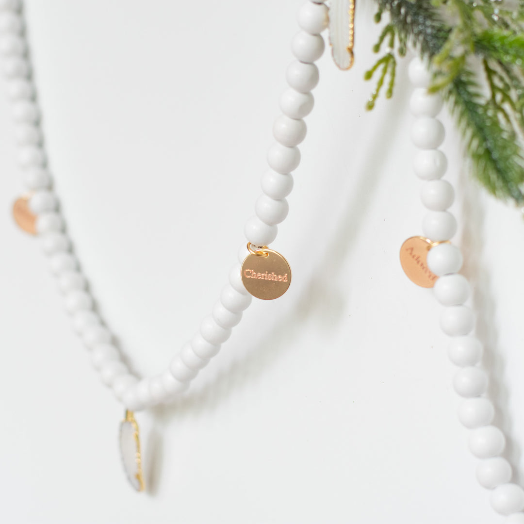 A close-up of a white garland with white feather charms and engraved gold charms highlighting the cherished charm and feather charm against a white background and pine branches.