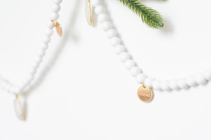 A close-up of a white garland with white feather charms and engraved gold charms highlighting the adorned charm against a white background and pine branches.