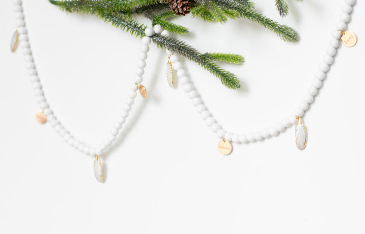 A wide shot of a white garland with white feather charms  and engraved gold charms hung up by pine branches against a white background