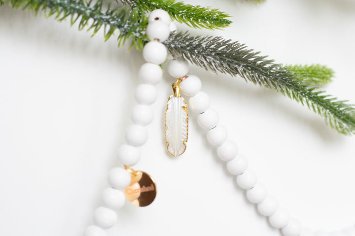 A close-up of a white garland with white feather charms and engraved gold charms against a white background with pine branches.