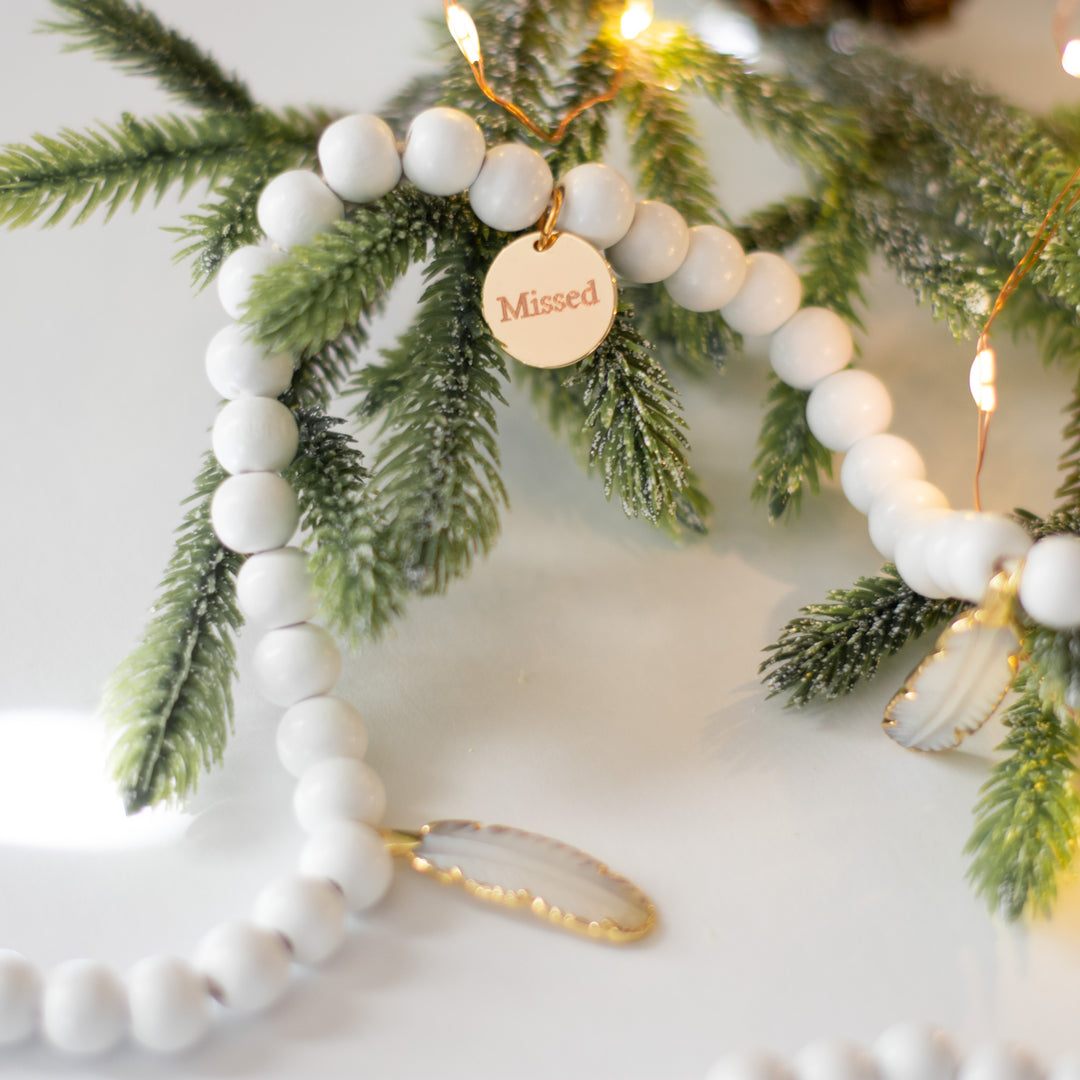 A close-up of a white garland with white feather charms and engraved gold charms against a white background and pine branches.