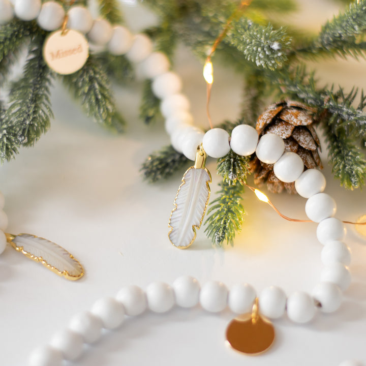 A close-up of a white garland with white feather charms and engraved gold charms against a white background with lights and pine branches.