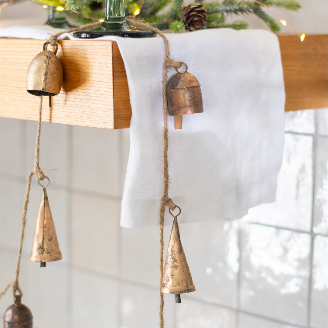 A close-up of a garland with gold bells resting on a shelf next to a pine branch and pine cone