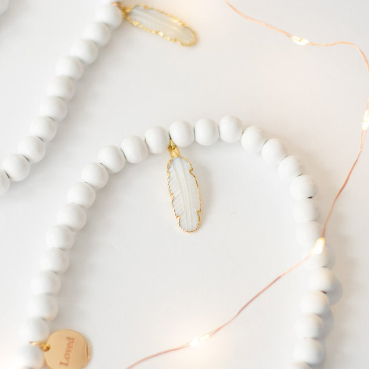 A close-up of a white garland with white feather charms and engraved gold charms against a white background with a string light.