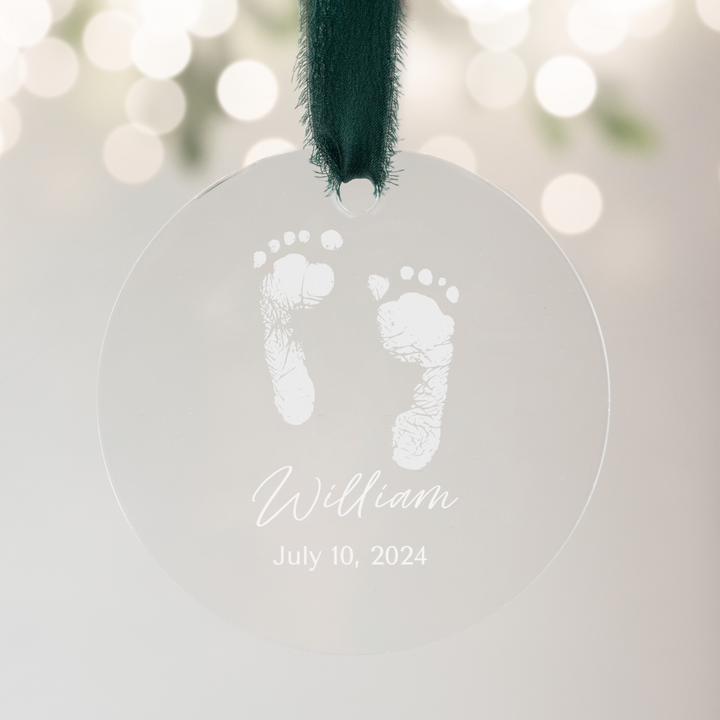 A close-up of a personalized acrylic glass ornament with a baby footprint, the text "William" and "July 10, 2024" engraved, and a dark green ribbon at the top with pine branches in the background.