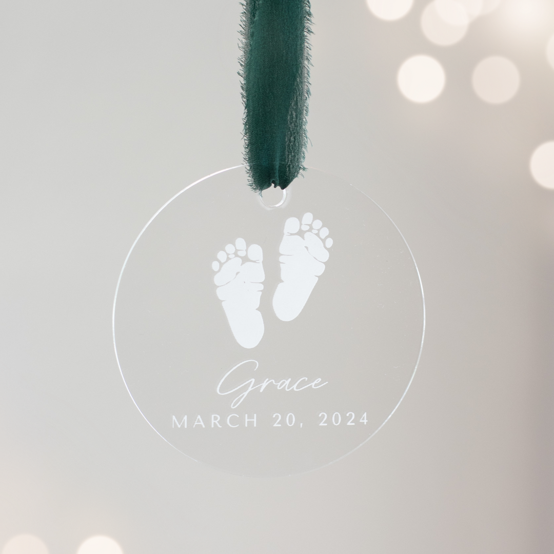 A close-up of a personalized acrylic glass ornament with a baby footprint, the text "Grace" and "March 20, 2024" engraved, and a dark green ribbon at the topwith lights in the background.