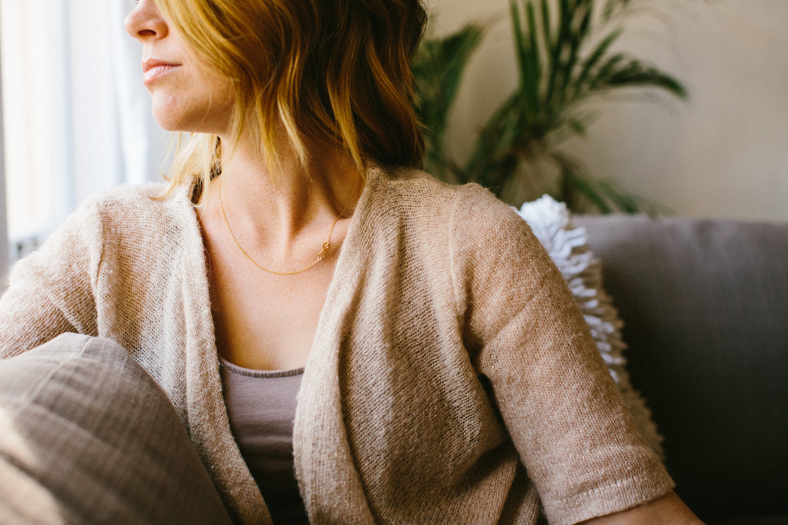 photo of a woman with blonde hair sitting on grey couch wearing a tan elbow length cardigan with a brown tank top underneath and  a gold necklace with an infinity symbol on it