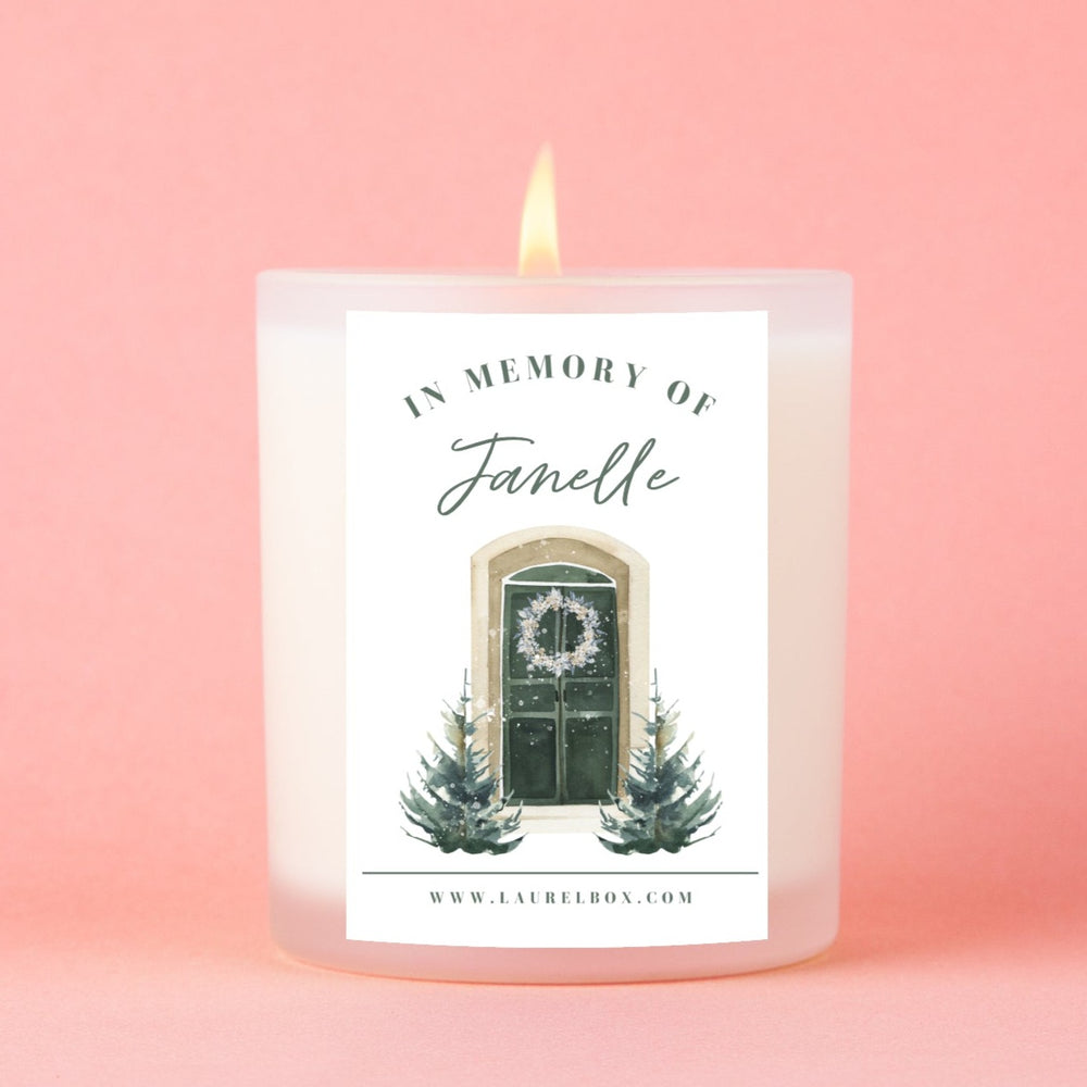 A white-coated candle with a white sticker with the text "In Memory of Janelle" and two Christmas trees and a door below the text against a pink background