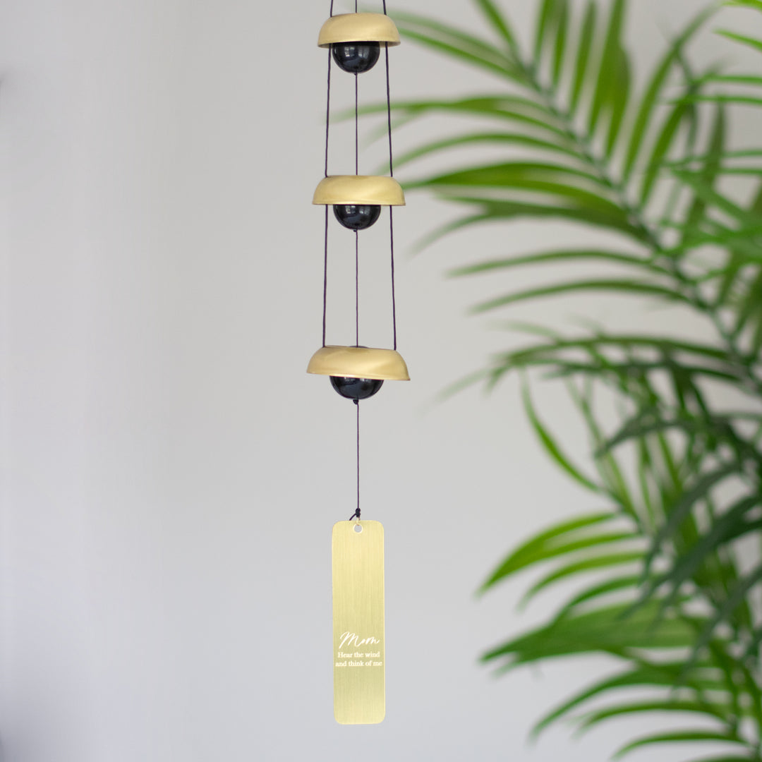 A wind chime hanging inside next to a plant that features three metallic bells, black cords, and a vertical rectangular tag at the bottom with the text mom and Hear the wind and think of me
