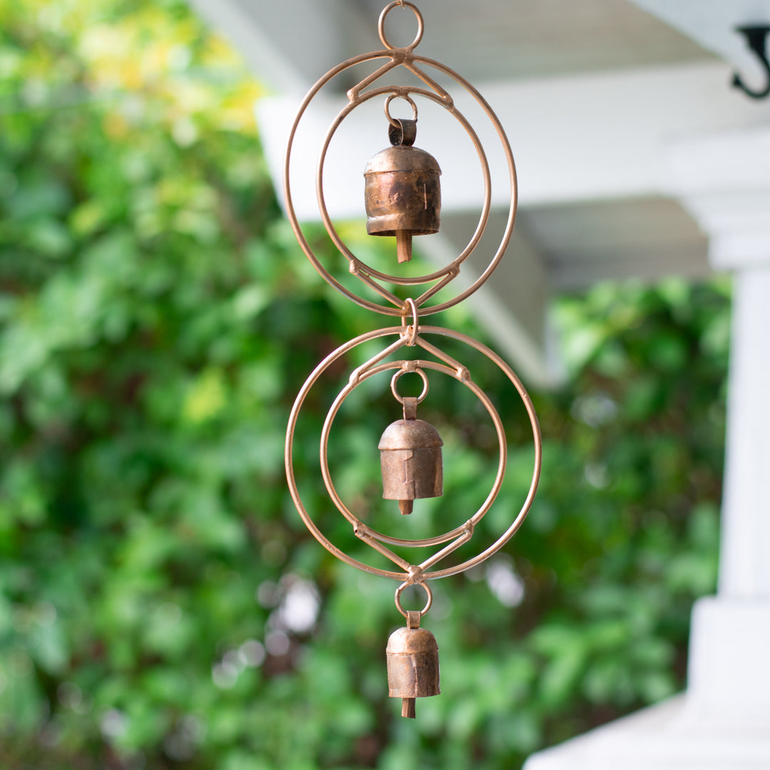 Close-up of a rustic wind chime of recycled tin and iron coated in copper showcasing three bells with a blurred background of a blurred bushes