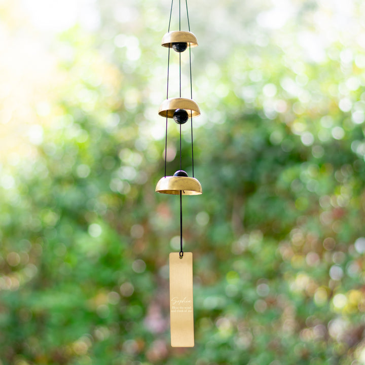 A wind chime hanging outside that features three metallic bells, black cords, and a vertical rectangular tag at the bottom with the text Sophia and Hear the wind and think of me