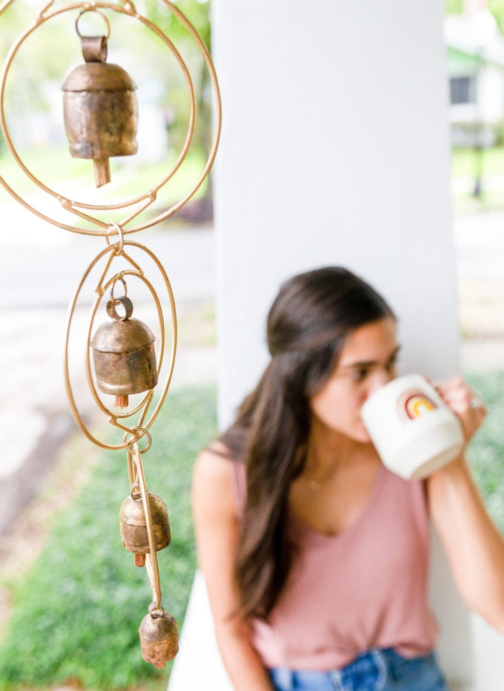 Close-up of a rustic wind chime of recycled tin and iron coated in copper with four bells and a woman blurred in the background drinking from a mug with a rainbow
