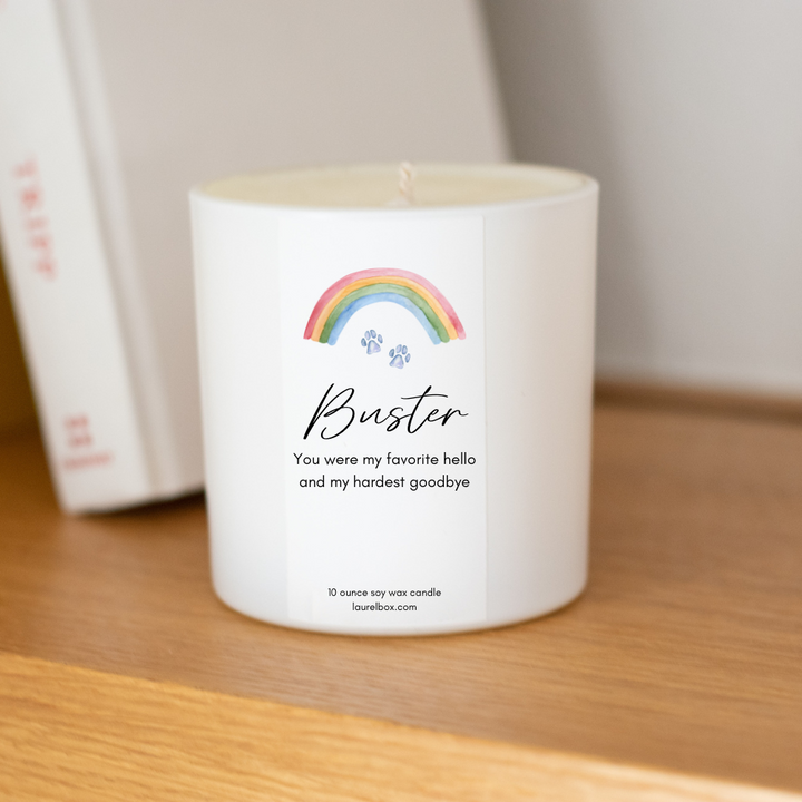 A white-coated candle with a white sticker including the text Bear You were my favorite hello and my hardest goodbye with a paw print and rainbow at the top. Sitting on a wooden surface with books in the background