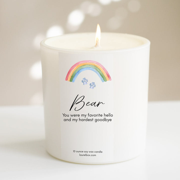 A white-coated candle with a white sticker including the text Bear You were my favorite hello and my hardest goodbye with a paw print and rainbow at the top. Sitting on a white surface.