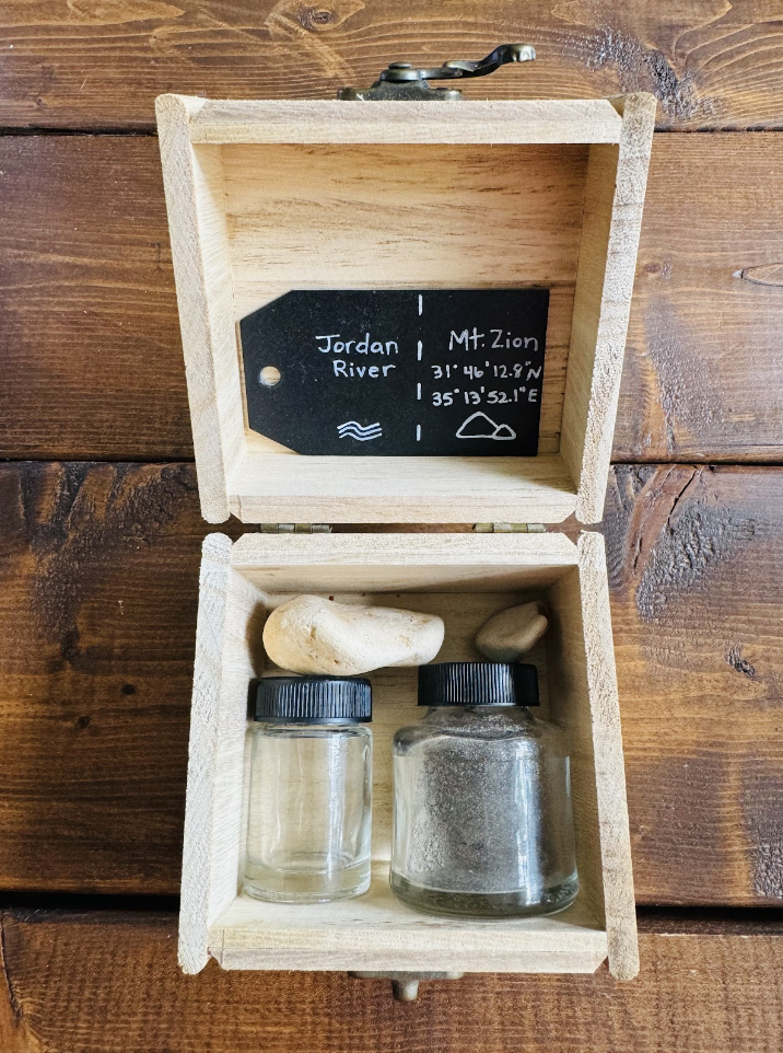 photo of the inside of a wooden box containing an empty jar with a black lid, a glass jar filled with a dark substance, a white rock and a small brown rock - with a black label in the lid with white wording
