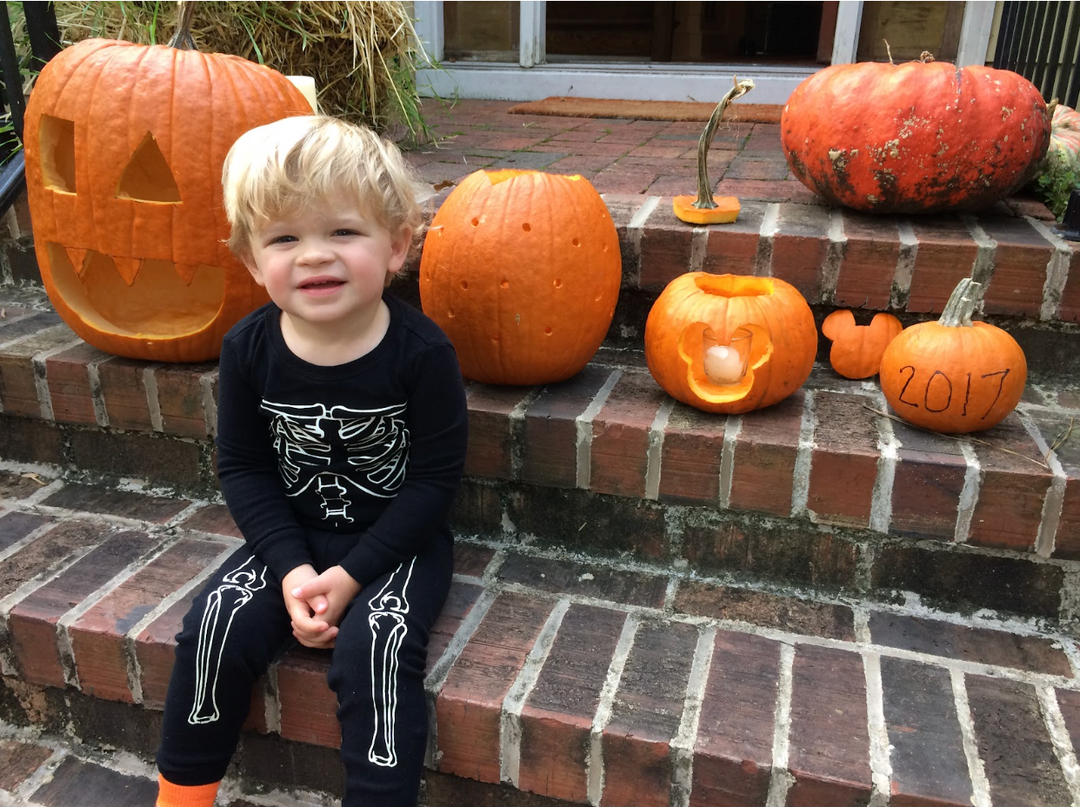 photo of a toddler with blonde short hair in a skeleton onesie sitting on a brick stoop surrounded by pumpkins