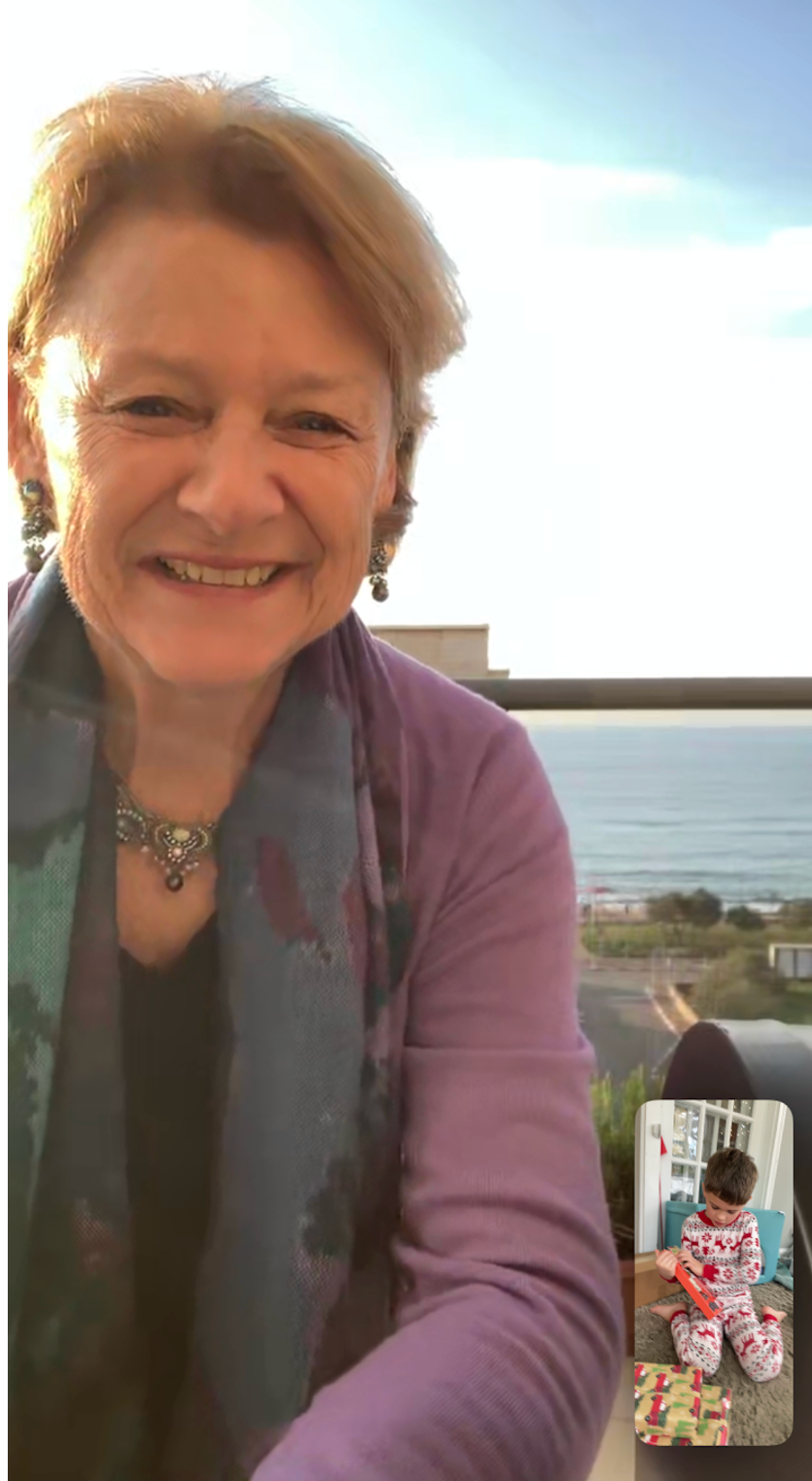 photo of an older woman wearing a purple shirt in front of a body of water smiling while face-timing with a young boy in Christmas pajamas opening gifts