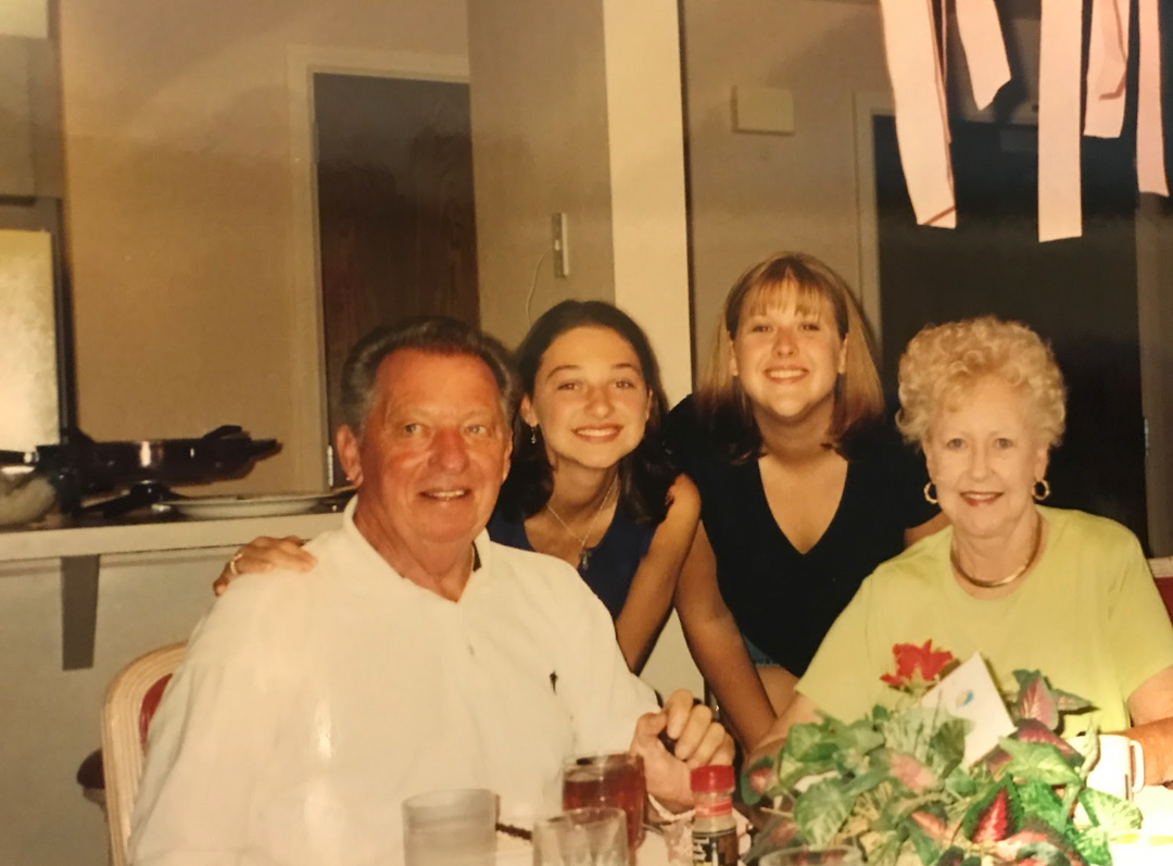 photo of two young women wearing black shirts crouching behind an older man in a white shirt and a woman in a yellow shirt who are seated at a table all smiling looking at the camera