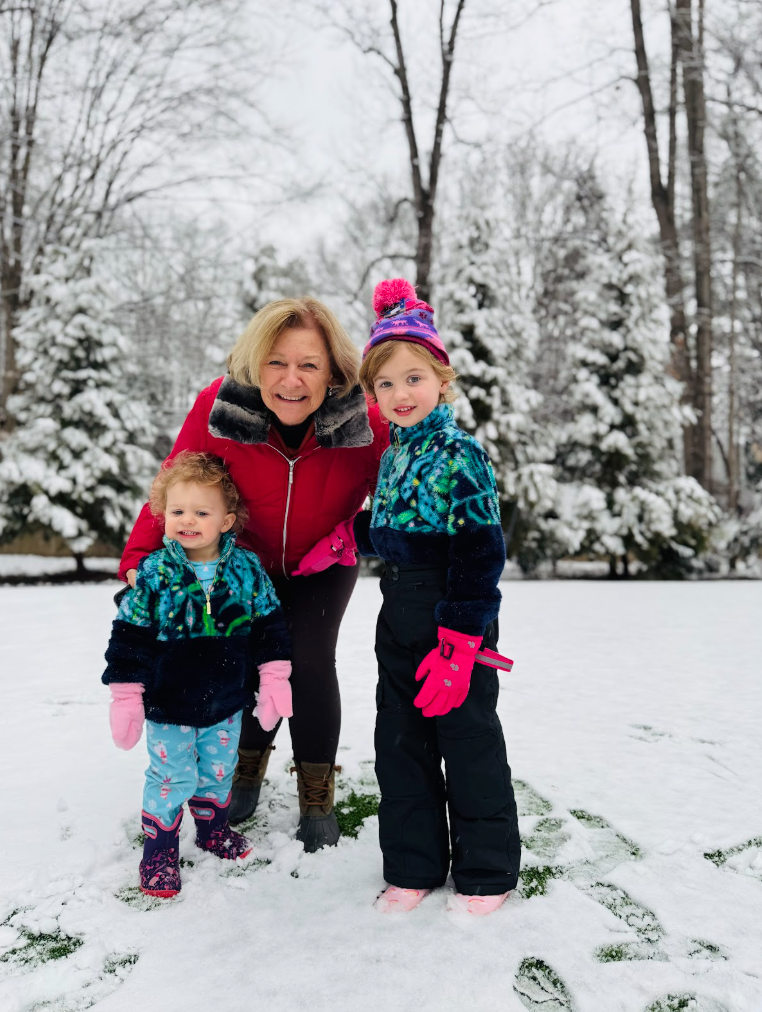 photo of a toddles in a blue, green and black snowsuit smiling next to a young child in the same snowsuit smiling next to an older woman wearing a red coat - all standing in a snow covered yard