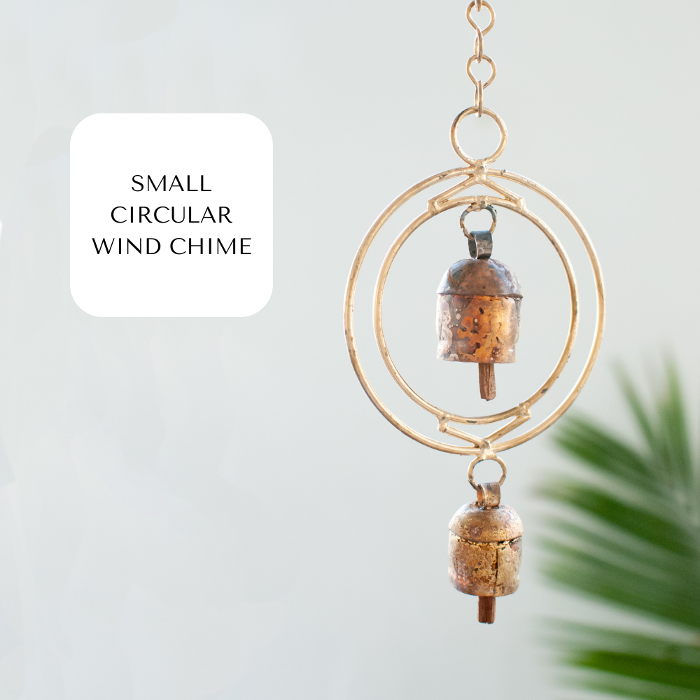 A close-up of a small rustic wind chime with two bells and golden metal circles against a white background and plant on the bottom right corner