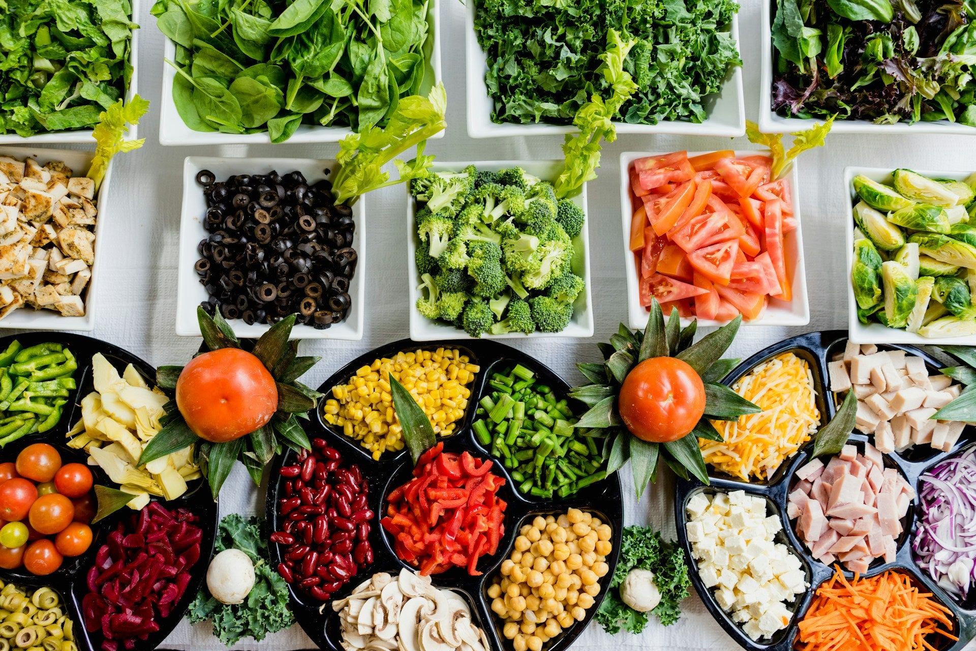 overhead photo of a table with bowls of lettuce, spinach, chicken, black olives, broccoli, tomatoes, Brussel sprouts with various vegetable trays in front of them