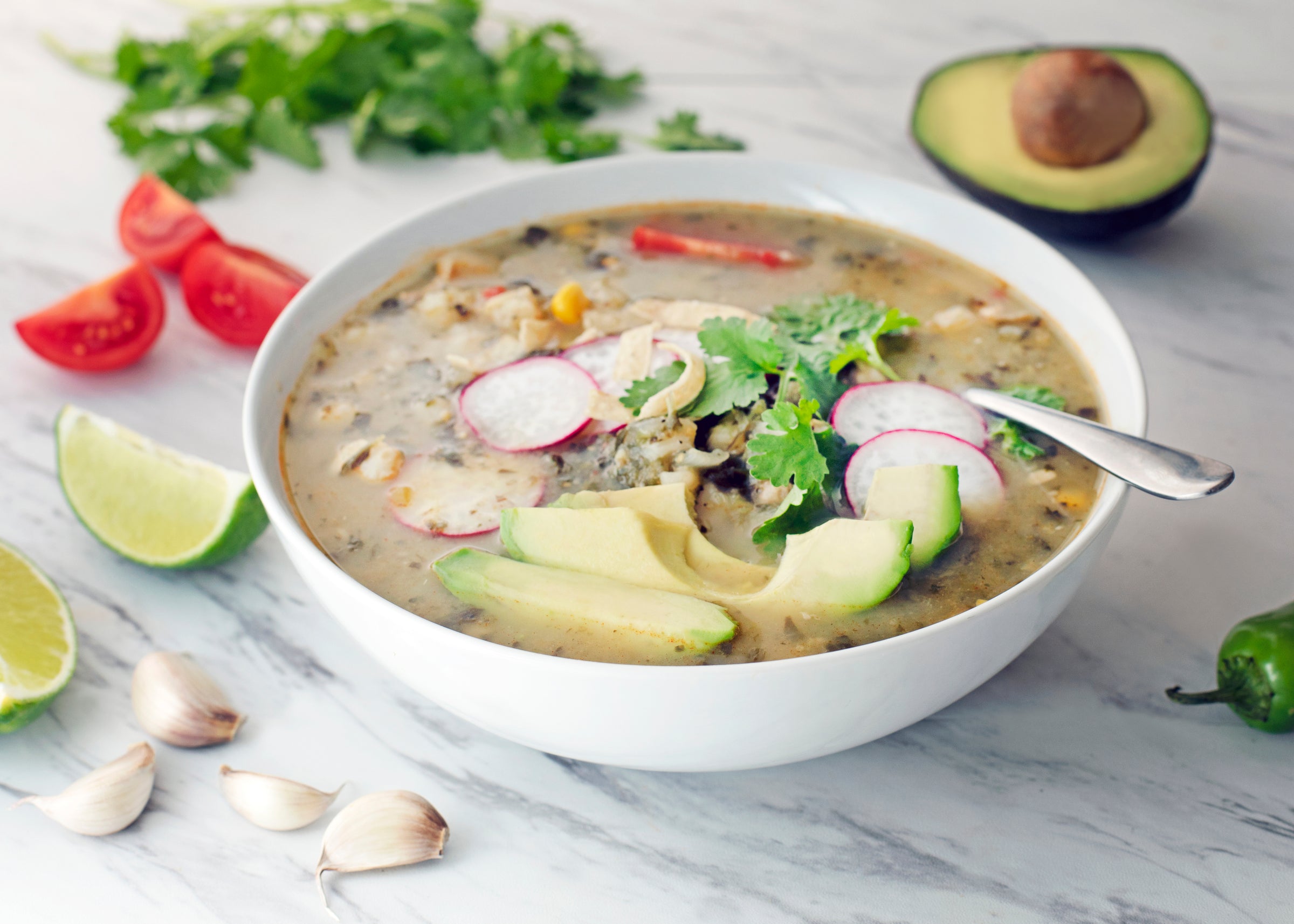 photo of a bowl of soup with radishes, avocado and other vegetables in it on a marble tabletop with tomatoes, limes, garlic, peppers and avocado surrounding the bowl