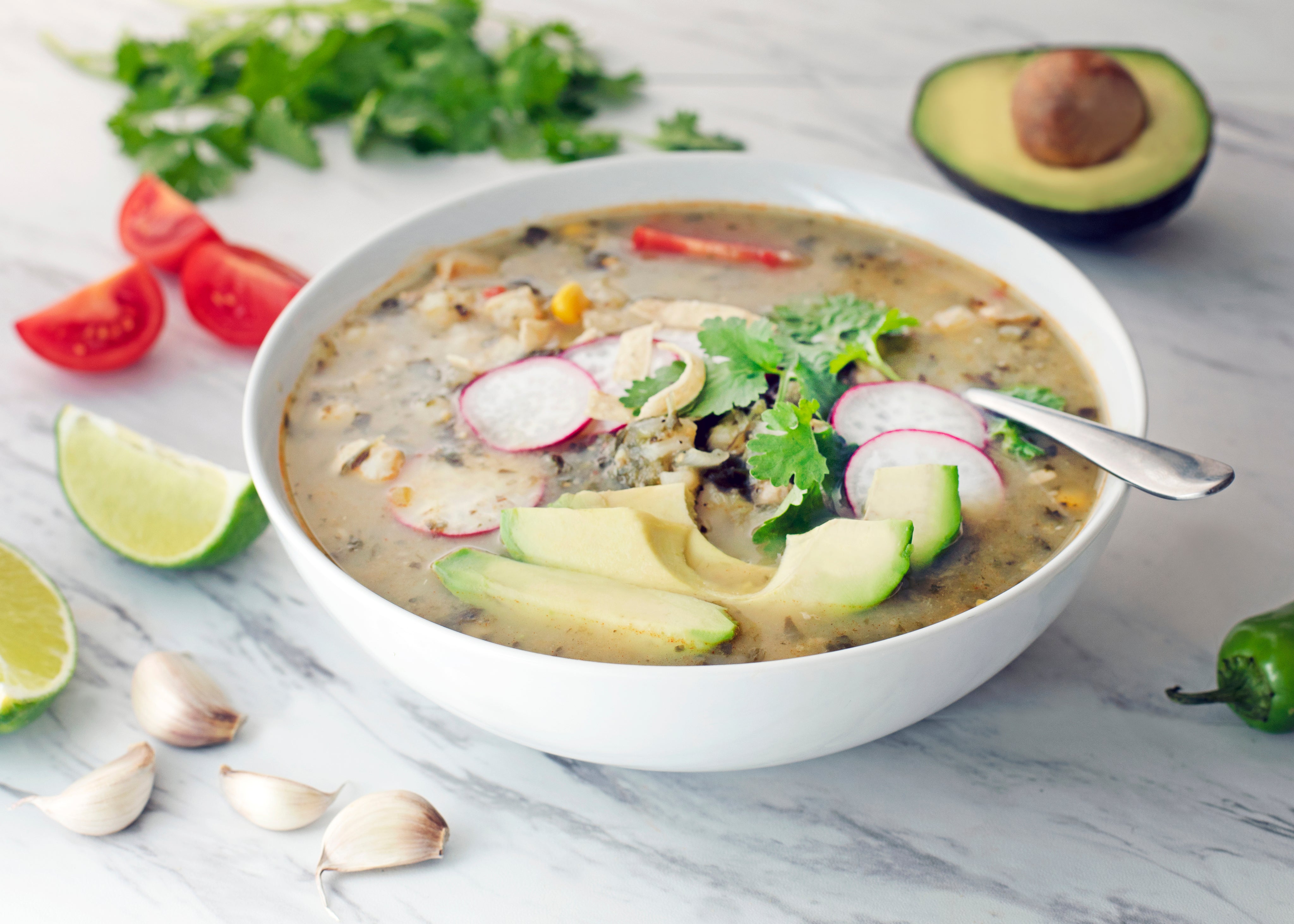 photo of a bowl of soup with radishes, avocado and other vegetables in it on a marble tabletop with tomatoes, limes, garlic, peppers and avocado surrounding the bowl