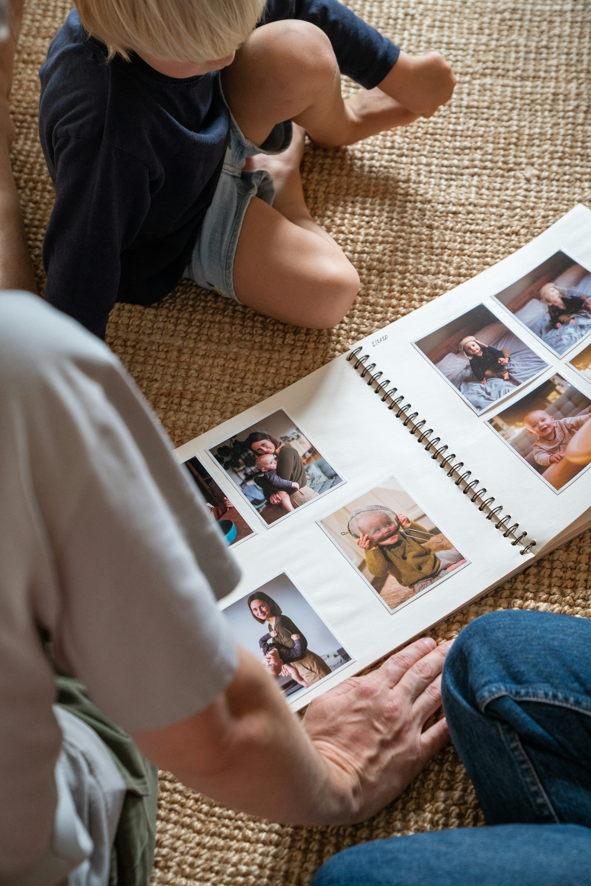 photo of a young boy with blonde hair wearing a blue outfit sat on a carpet with a home made photo book open with the hand of an older person nearby