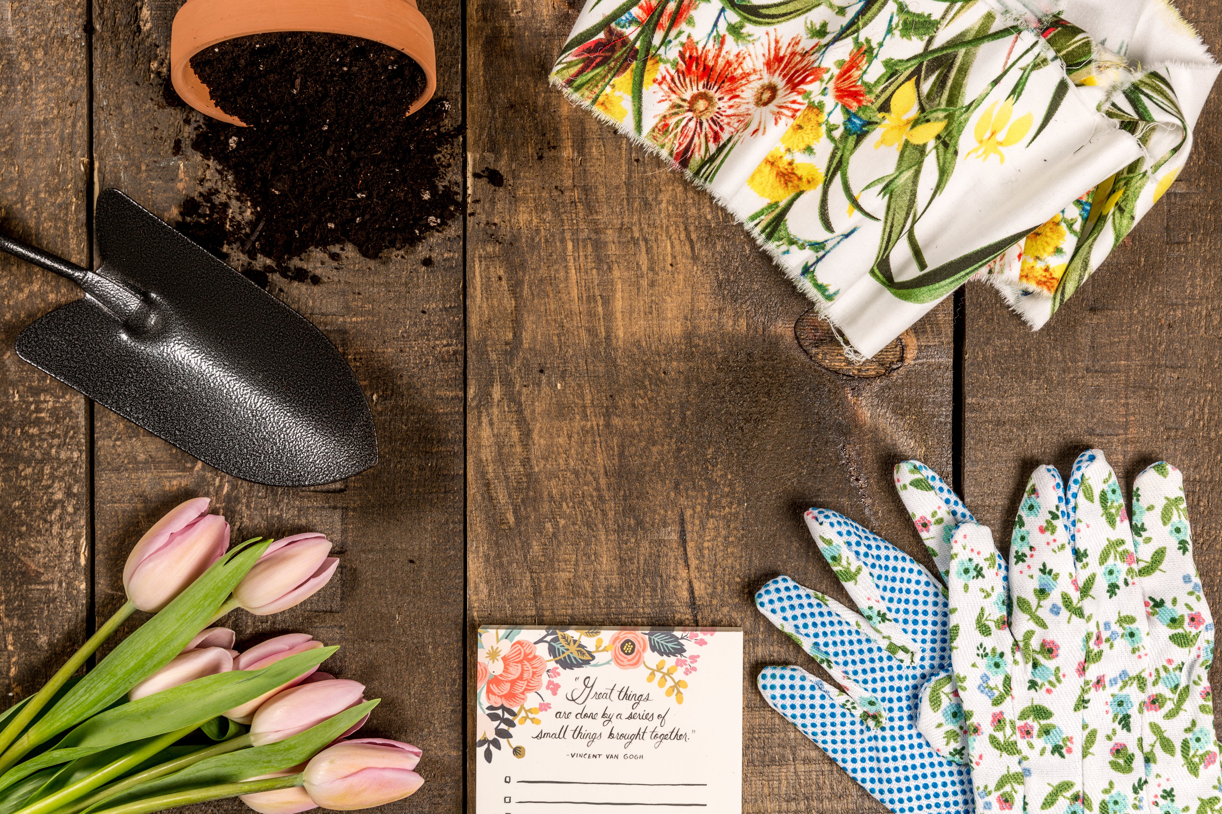 gardening tools - small shovel, terracotta pot with soil spilling out, towels with red, yellow & green flows on it, gardening gloves with a flower. pattern, a piece of paper with flowers at the top next to pink tulips - on top of dark wood planks