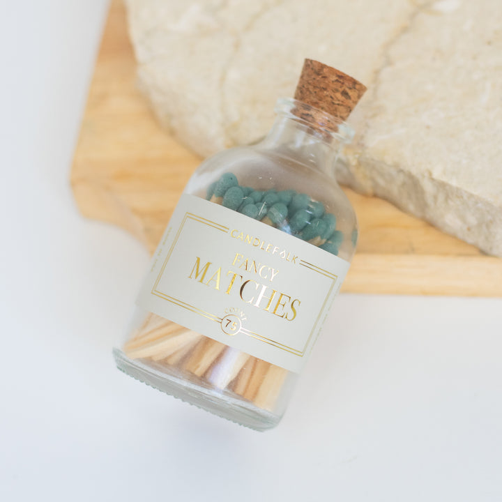 A glass container with a white label including Fancy Matches text filled with green matches angled to the right against a piece of granite and wood