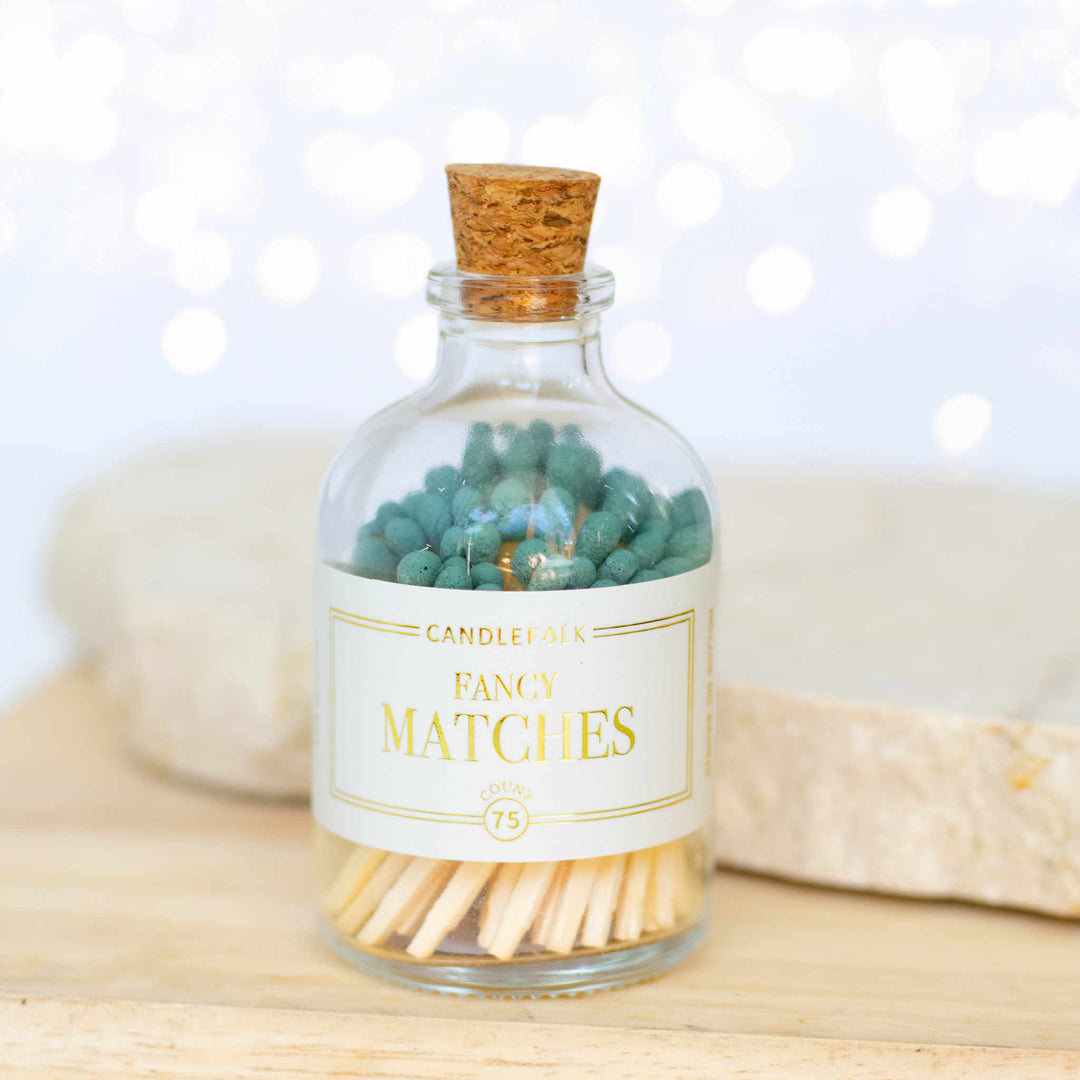 A glass container with a white label including Fancy Matches text filled with green matches sitting on a wooden table