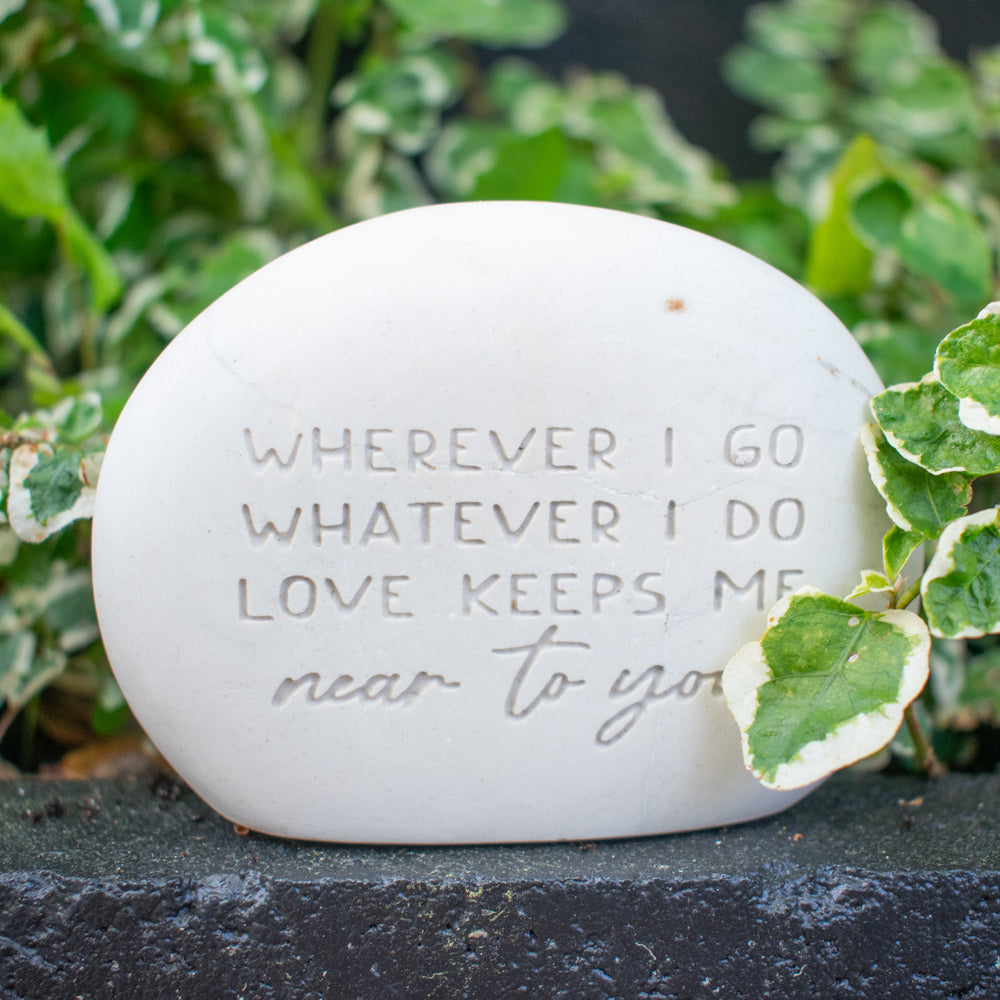 A memorial gemstone with Whenever I Go Whatever I Do Love Keeps Me Near To You engraved on it sitting outside on black stone and plants in the background